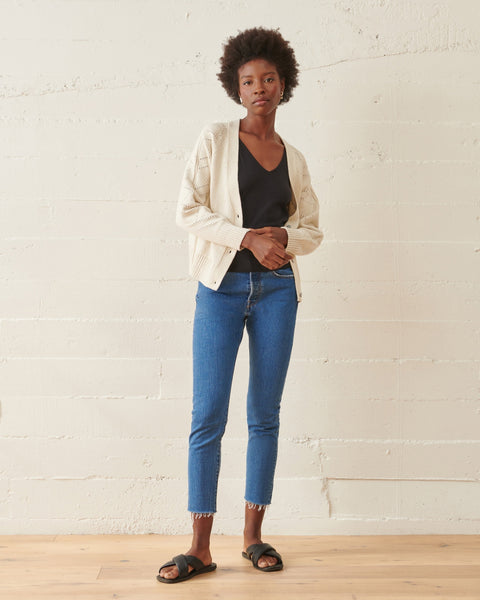 woman stands with  hudson cardigan, worn open,  layered over dark tank top. She's holding one cuff with her opposite hand, a soft posture, a relaxed pose, for a relaxed outfit.  She's wearing  skinny denim, with a distressed hem and sandals, a neutral background n light wooden floor